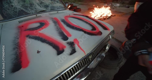Riot Fire. Protest. Vandalism Punk protester girl uses a red pressure paint finishing vandal graffiti riot ugly lettering on viciously crushed old car on the street