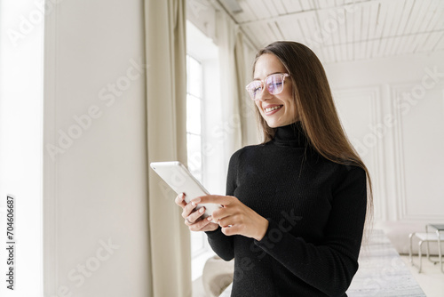 Elegant woman in black turtleneck using a tablet, standing by a window in a minimalist white room.