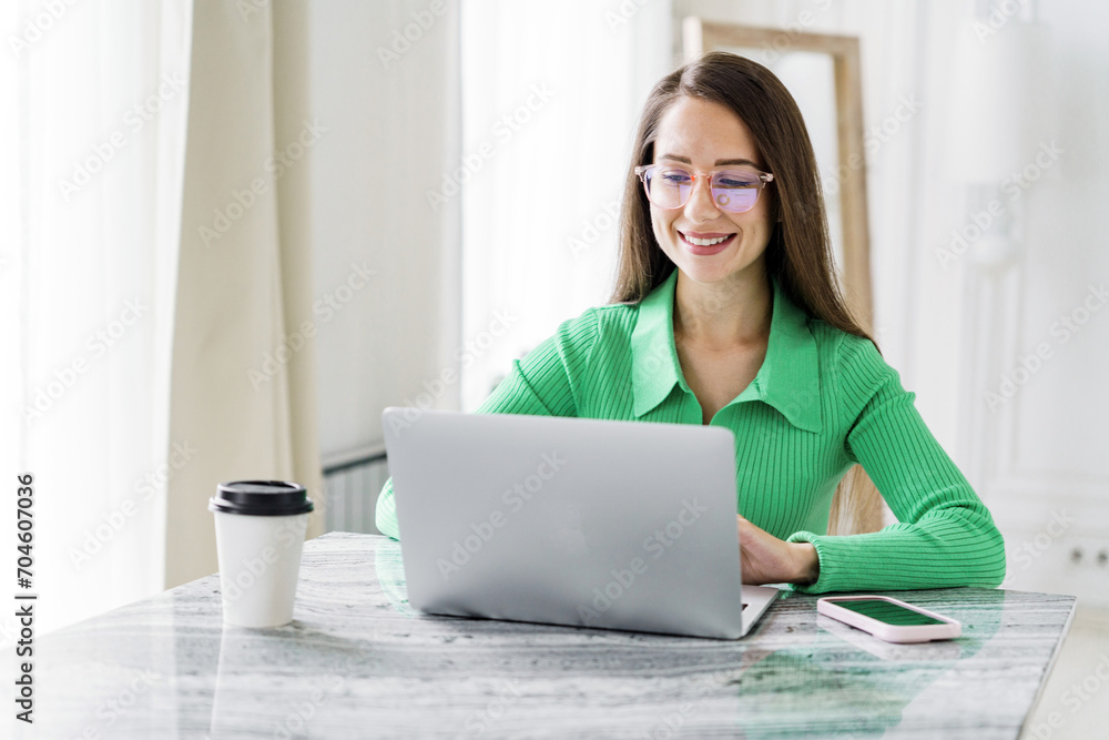 Efficient young woman with a laptop and coffee, engaged in work at a marble table in a well-lit room.
