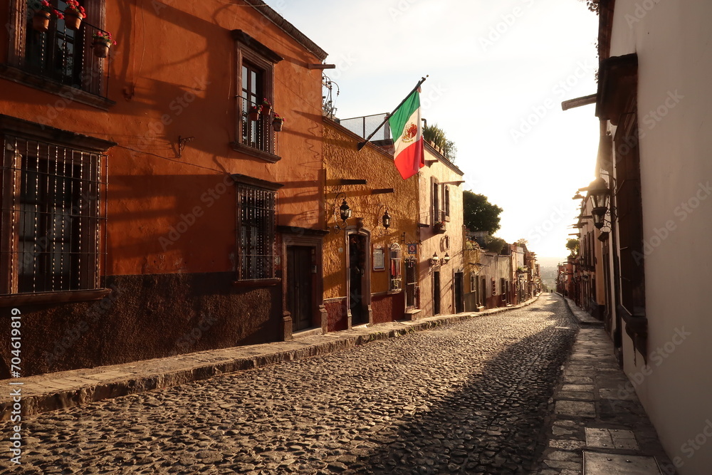 Obraz premium Typical cobblestone street in San Miguel de Allende at golden hour/sunset, a mexican flag is hanging on a facade, Mexico