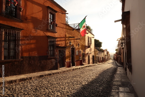 Typical cobblestone street in San Miguel de Allende at golden hour/sunset, a mexican flag is hanging on a facade, Mexico