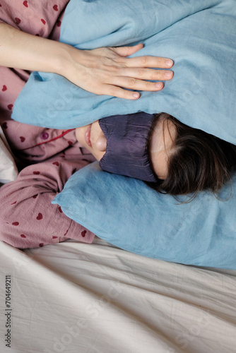 young woman in bed wakes up, insomnia, sleep disorder. person in sleep mask holds onto pillows, covers his ears, face, has headache in morning. lady in pajamas can't sleep, noise neighbors from street © MyJuly