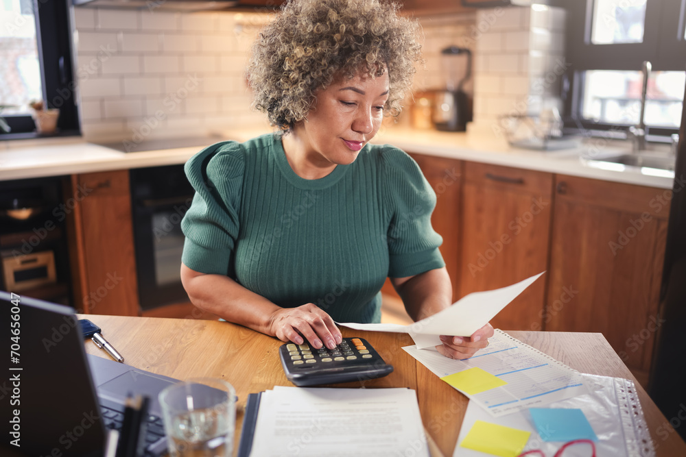 A mature entrepreneur woman of mixed race working on a computer at home. Woman small business owner. Woman working on a computer, calculating monthly expenses, mortgage payments, doing home budgeting A mature entrepreneur woman of mixed race working on a computer at home. Woman small business owner. Woman working on a computer, calculating monthly expenses, mortgage payments, doing home budgeting