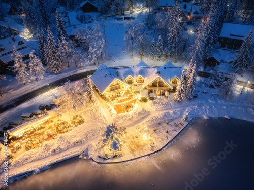 Aerial view of illuminated houses in fairy village in snow, forest, Jasna lake, street lights at winter night. Top view of alpine countryside, snowy pine trees at dusk. Kranjska Gora, Slovenia