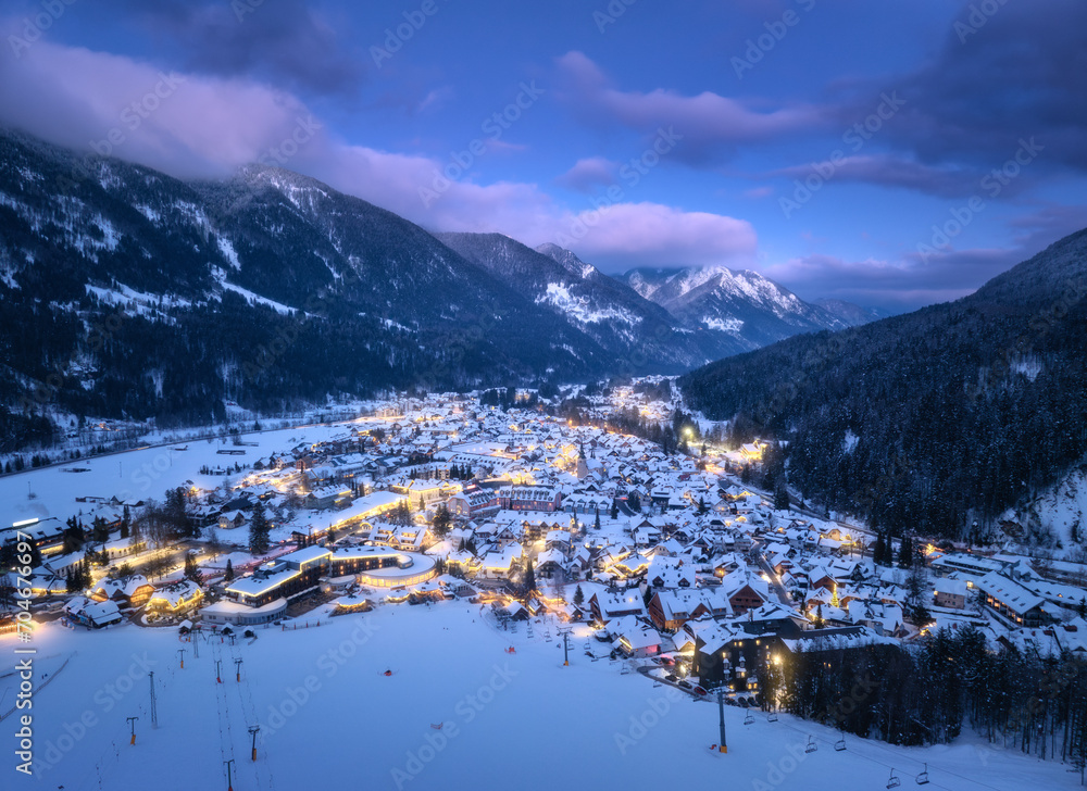 Aerial view of beautiful alpine village at cold winter night. Kranjska Gora, Slovenia. Top drone view of buildings in snow, street lights, road, snowy alpine mountains, sky at dusk. Triglav park