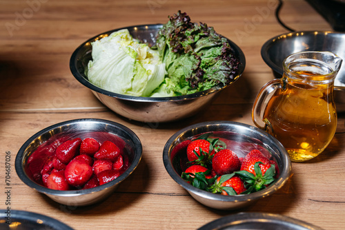 Bowls with raw cooking ingredients, a bowl of fresh strawberries, a bowl of lettuce leaves, a pitcher of oil on a wooden table surface