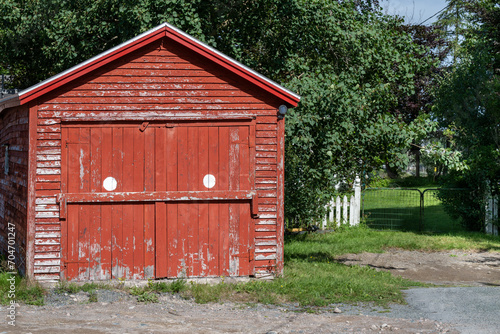 A vintage vibrant red wooden garage or barn. There are two wood doors with white spots, a peaked roof, and peeling paint. The outcrop structure is near large maple trees and a white picket fence. 