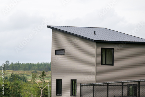 The exterior corner of a modern wooden tiny house. It has grey vinyl walls with black trim, closed glass windows, slanted flat roof, black metal backyard fence and lush green views of the countryside.