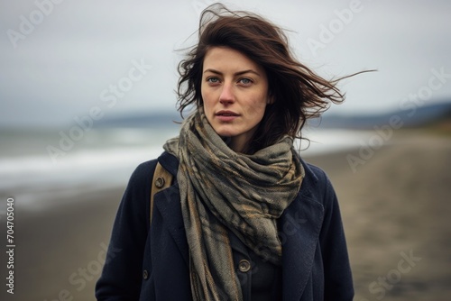 A woman stands on the beach in cloudy, windy weather wrapped in a scarf and coat