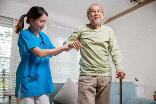 International Day for the Elderly. Young carer supporting helping senior man stand up with walking stick from sofa, Nurse wearing blue uniform help helping his patient senior old man with walking cane