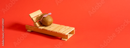 Kiwi with sunglasses resting on the wooden beach bed. Summer vacation concept on red background