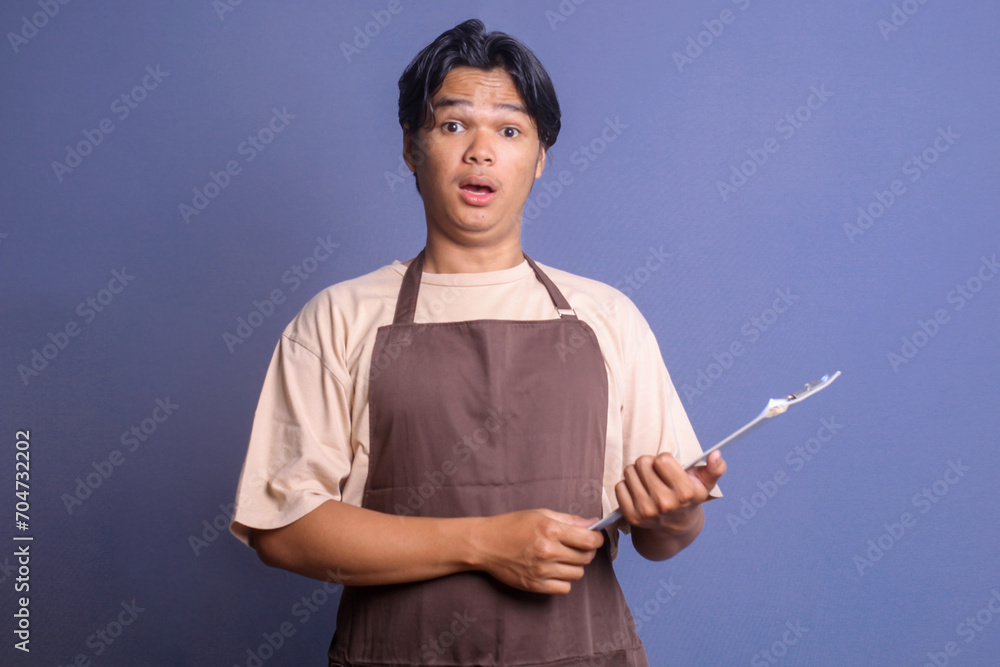 Young barista in brown apron holding clipboard and looking at camera with shocked surprise expression Young barista in brown apron holding clipboard and looking at camera with shocked surprise expression