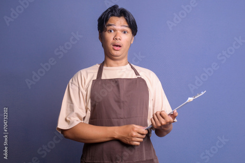Young barista in brown apron holding clipboard and looking at camera with shocked surprise expression