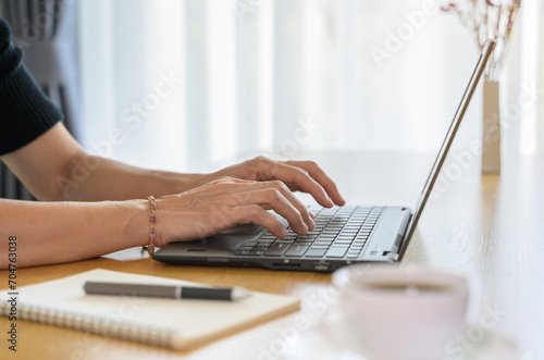 Close up on hands of female in causal cloth working on laptop computer at home while sitting at desk with coffee cup and notebook 