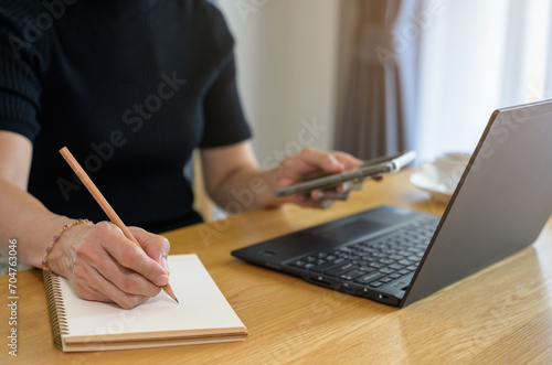 Freelancer or businesswoman working on smart phone and writing on paper notebook at home office while sitting in front of laptop computer. Online working from home concept. Close up of woman hand taki