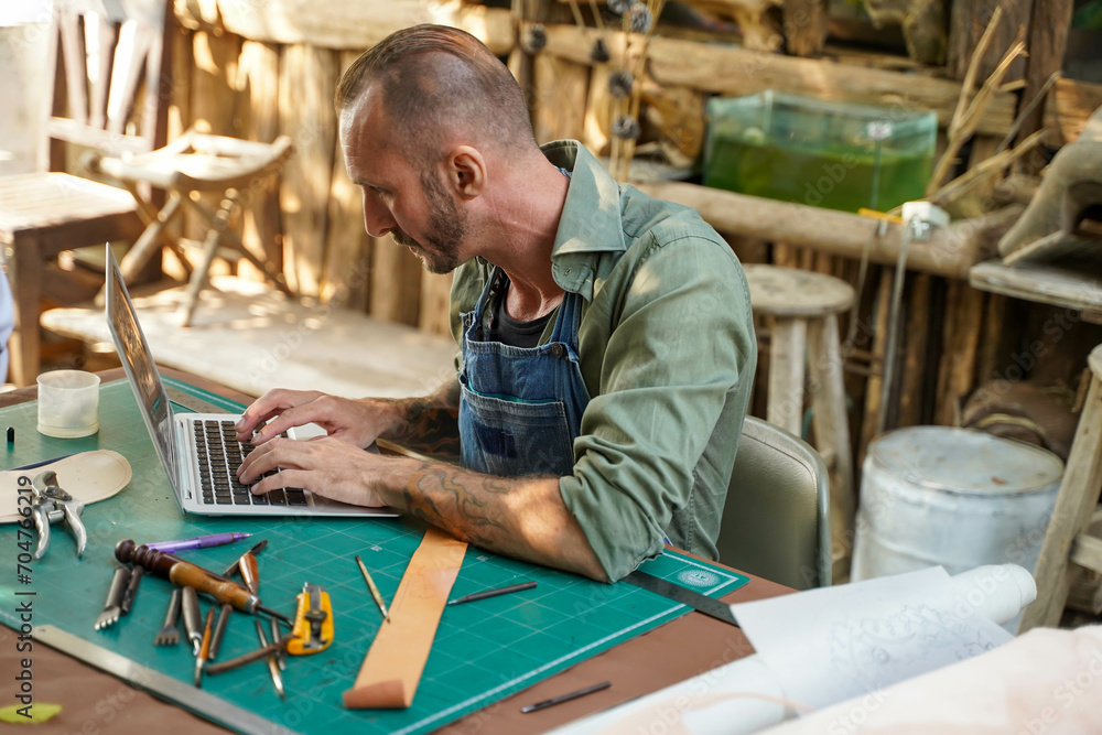 Closeup leather craftsman using a laptop check the leather rolls in the stock with customer demand in the work shop. Closeup leather craftsman using a laptop check the leather rolls in the stock with customer demand in the work shop.