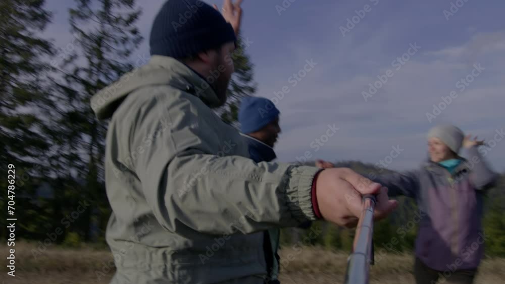 POV from smartphone front camera. Caucasian man with selfie stick records video with multiethnic couple on top of hill and dances with them. Group of diverse hiking buddies during trip in mountains. POV from smartphone front camera. Caucasian man with selfie stick records video with multiethnic couple on top of hill and dances with them. Group of diverse hiking buddies during trip in mountains.