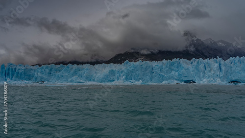 A wall of blue ice with cracks and sharp peaks stretches over a turquoise lake. Melted ice floes float in the water. The mountains are shrouded in clouds and fog. Perito Moreno glacier. El Calafate. 