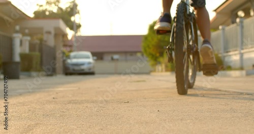 Slow motion low angle shot while a 9 year old Asian boy rides a bicycle in the evening on a weekend in an urban village and there is a beautiful golden sunlight in the scene.