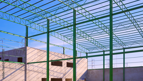 Green metal columns and roof beam outline with concrete industrial office building structure in new factory construction site area against blue sky background, low angle and perspective side view