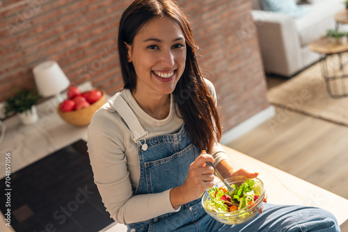 Beauty smiling woman eating healthy salad while sitting on the kitchen table at home.