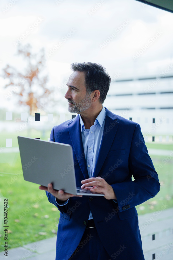 Contemplative businessman holding laptop and looking through glass window in office Contemplative businessman holding laptop and looking through glass window in office