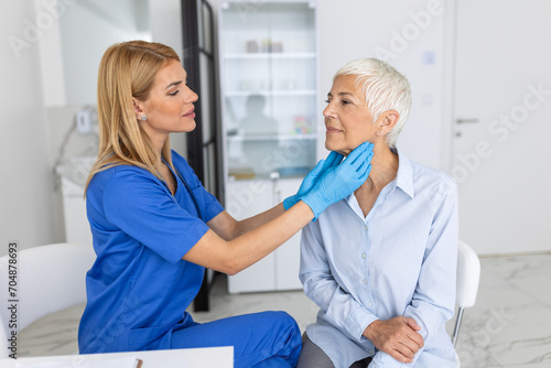 Friendly woman doctor wearing gloves checking sore throat or thyroid glands, touching neck of senior female patient visiting clinic office. Thyroid cancer prevention concept photo