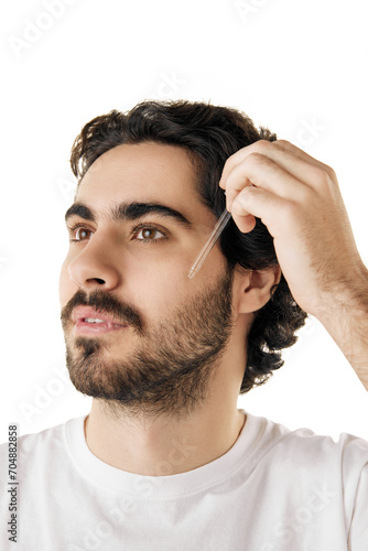 Portrait of handsome man holds jar of cosmetic product applying with pipette beard oil or facial serum against white studio background. Concept of male facial care routine, cosmetic product.