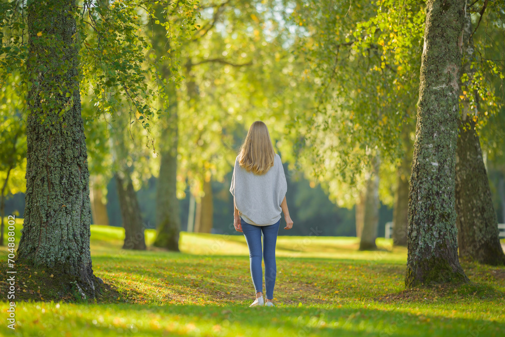 Young adult blonde woman slowly walking on green grass at city park in beautiful warm sunny autumn day. Spending time alone and enjoying freedom at nature. Peaceful atmosphere. Back view. Young adult blonde woman slowly walking on green grass at city park in beautiful warm sunny autumn day. Spending time alone and enjoying freedom at nature. Peaceful atmosphere. Back view.