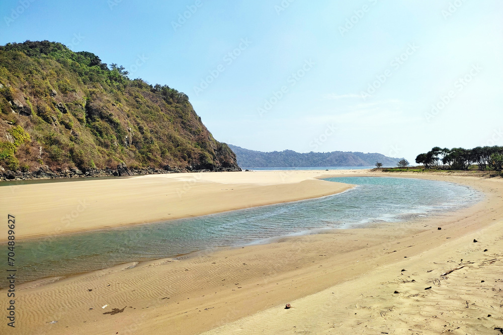 Muara Mbaduk Beach in daytime, receding sea, calm river flowing to the Indian Ocean. Around it, white sand stretches and coral hills with vegetation.