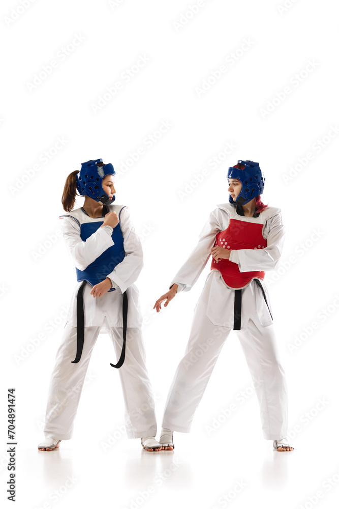 Full-length image of young competitive girls in dobok and helmet, practicing taekwondo, training isolated over white background. Concept of martial arts, combat sport, competition, action Full-length image of young competitive girls in dobok and helmet, practicing taekwondo, training isolated over white background. Concept of martial arts, combat sport, competition, action