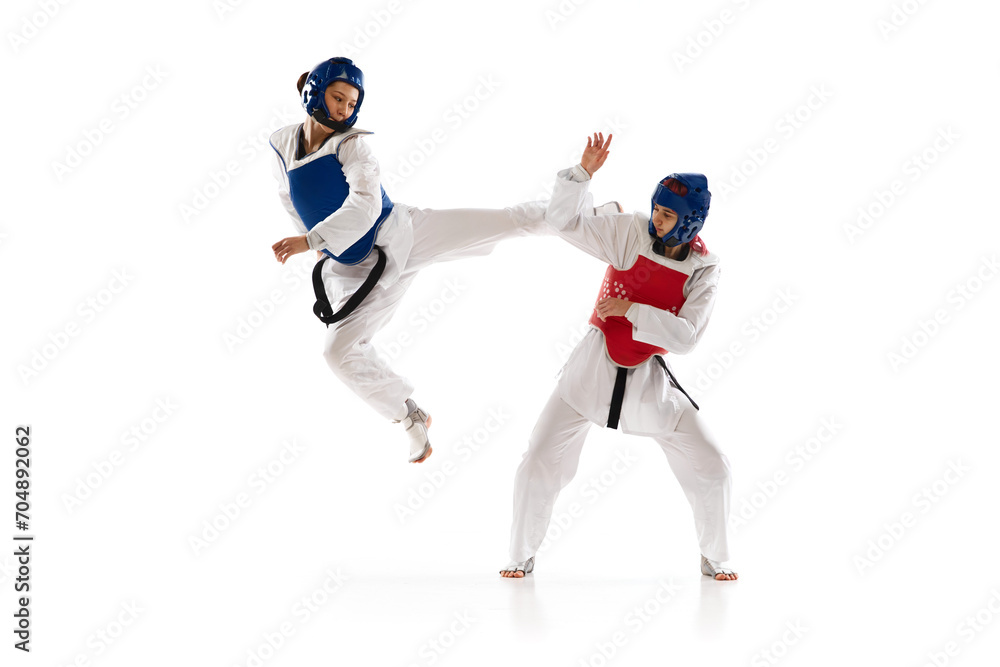 Dynamic image of young girls in helmet and dobok practicing taekwondo stunts, fighting isolated over white background. Concept of martial arts, combat sport, competition, action
