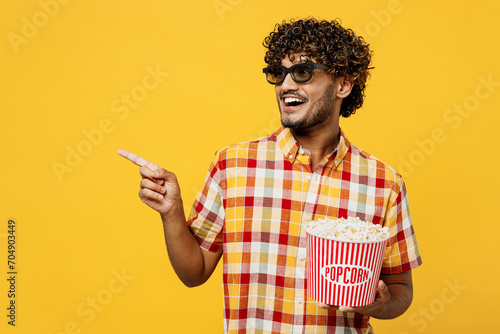 Young smiling happy cool Indian man wear 3d glasses watch movie film hold bucket of popcorn in cinema point index finger aside on area copy space isolated on plain yellow background studio portrait.