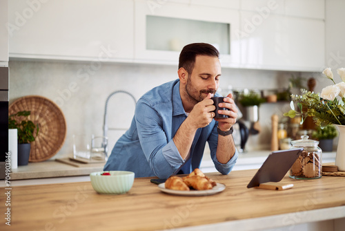 A content freelance eyes-closed man holding a cup of coffee in his hands while leaning on a kitchen counter in front a tablet.