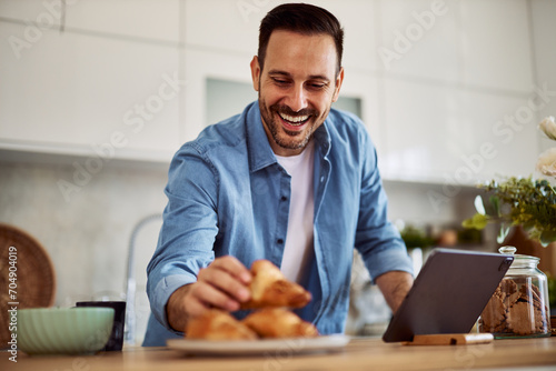 A happy freelance man leaning forward to take a croissant while standing in front of a tablet. photo