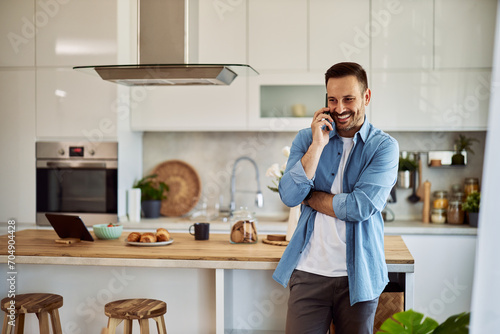 A smiling young adult man booking a hotel room for a vacation over a phone call while leaning against a kitchen counter.