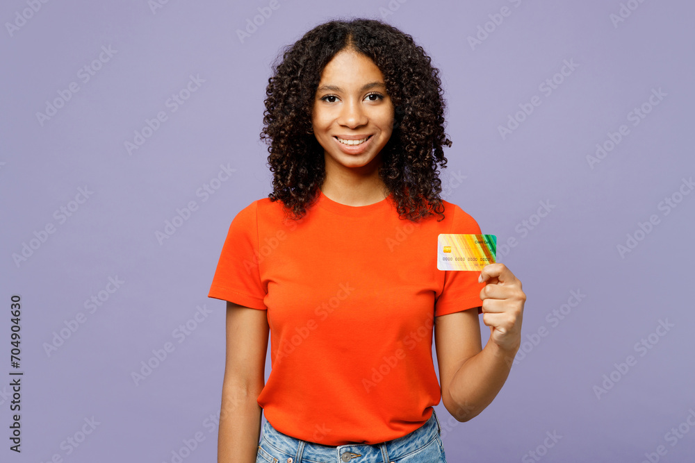 Little fun kid teen girl of African American ethnicity wear orange t-shirt hold in hand mock up of credit bank card isolated on plain pastel light purple background studio Childhood lifestyle concept. Little fun kid teen girl of African American ethnicity wear orange t-shirt hold in hand mock up of credit bank card isolated on plain pastel light purple background studio Childhood lifestyle concept.