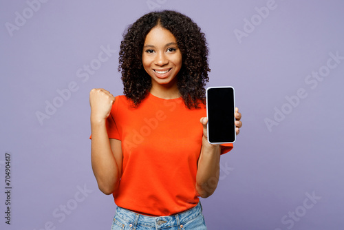 Little kid teen girl of African American ethnicity wear orange t-shirt hold use blank screen area mobile cell phone do winner gesture isolated on plain purple background. Childhood lifestyle concept.