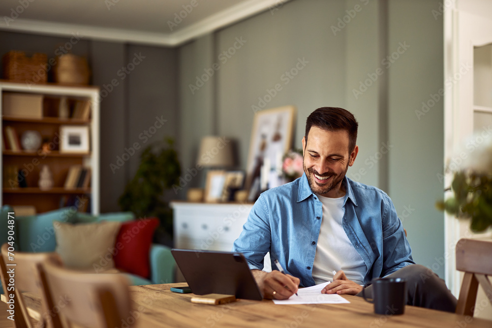 A cheerful young adult man interviewing a candidate over an online platform on a tablet while writing notes on a sheet of paper in front of him. - obrazy, fototapety, plakaty 