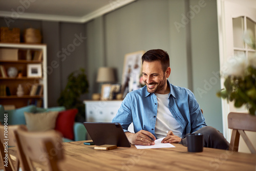 A happy young adult man conducting an online interview on a tablet holding a pen and a sheet of paper in front of him.