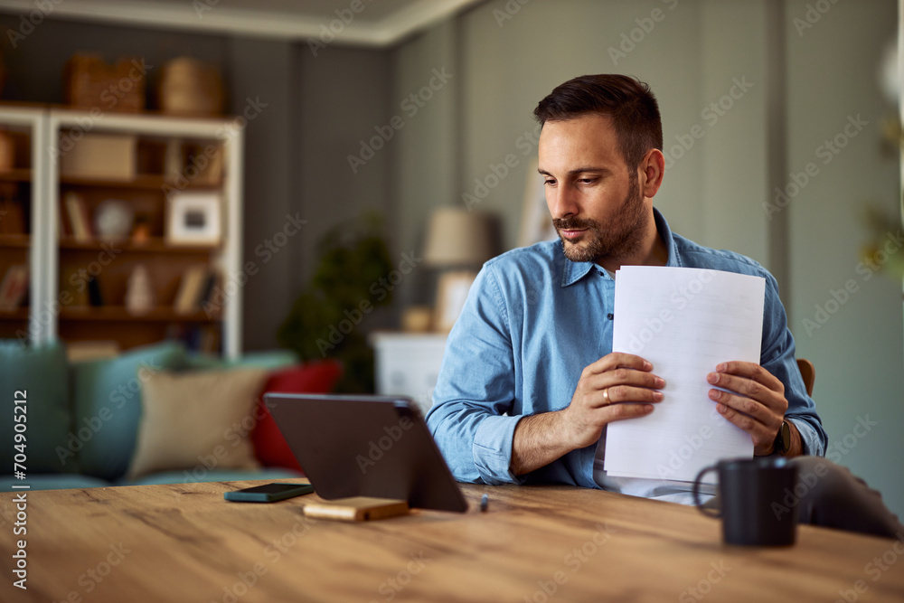 A serious young adult man organizing documents while looking at a tablet for work. A serious young adult man organizing documents while looking at a tablet for work.