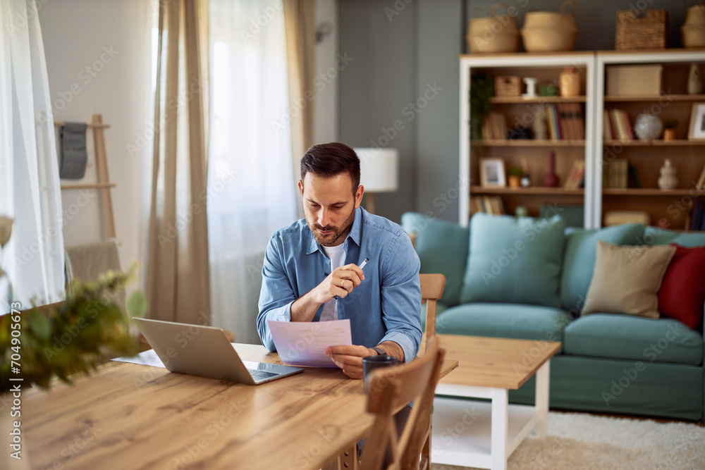 A focused male remote worker analyzing data on a paper he is holding while sitting at a desk in front of a laptop. A focused male remote worker analyzing data on a paper he is holding while sitting at a desk in front of a laptop.