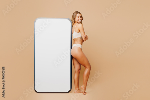 Full body young lady woman with slim body perfect skin wear nude top bra lingerie stand near big huge blank screen area mobile cell phone isolated on plain beige background Lifestyle diet fit concept photo