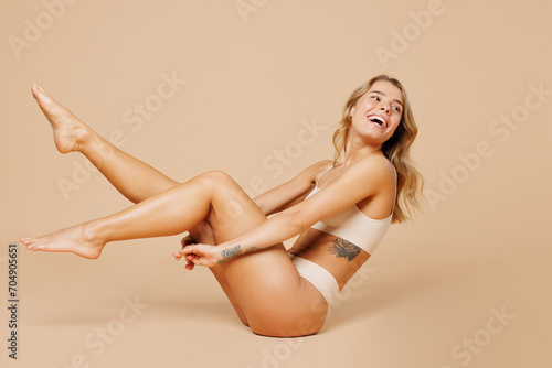Full body side view young nice lady woman with slim body perfect skin wearing nude top bra lingerie looking aside raise up legs isolated on plain pastel beige background. Lifestyle diet fit concept.