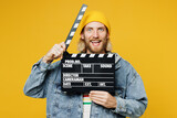Young cheerful happy man he wear denim shirt hoody beanie hat casual clothes hold in hand classic black film making clapperboard isolated on plain yellow background studio portrait. Lifestyle concept