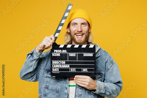 Young cheerful happy man he wear denim shirt hoody beanie hat casual clothes hold in hand classic black film making clapperboard isolated on plain yellow background studio portrait. Lifestyle concept photo