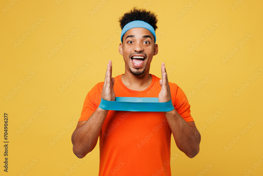 Young surprised fitness trainer instructor sporty man sportsman wear orange t-shirt use elastic band on hands spend time in home gym isolated on plain yellow background. Workout sport fit abs concept. Young surprised fitness trainer instructor sporty man sportsman wear orange t-shirt use elastic band on hands spend time in home gym isolated on plain yellow background. Workout sport fit abs concept.