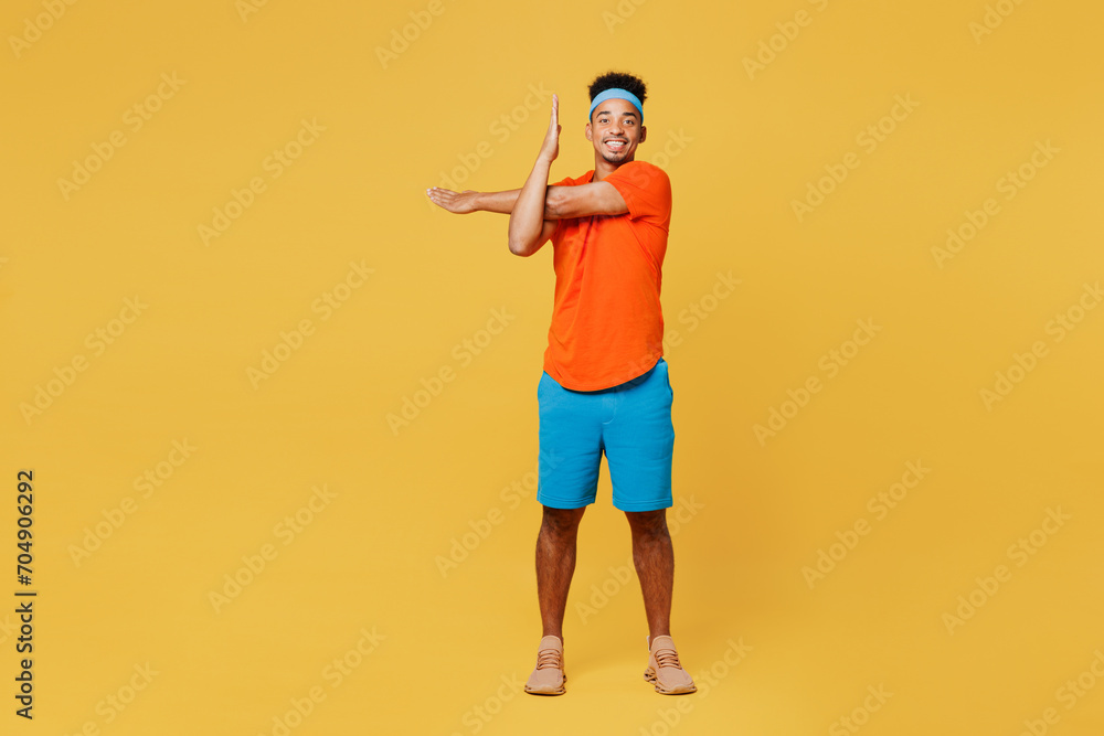 Fototapeta premium Full body happy young fitness trainer sporty man sportsman wear orange t-shirt do stretch exercise for hand spend time in home gym isolated on plain yellow background. Workout sport fit abs concept.