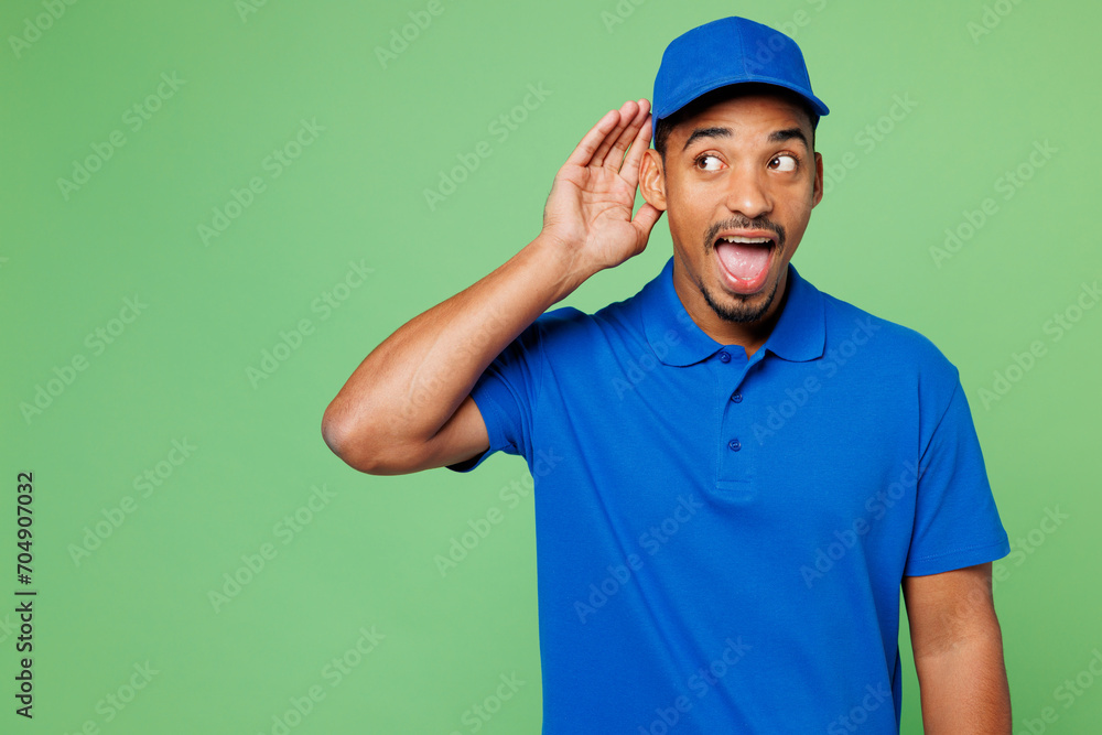 Professional delivery guy employee man wears blue cap t-shirt uniform workwear work as dealer courier try to hear you overhear listening intently isolated on plain green background. Service concept.