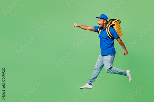 Full body delivery guy employee man wearing blue cap t-shirt uniform workwear yellow thermal food bag backpack work as dealer courier jump high run isolated on plain green background. Service concept.